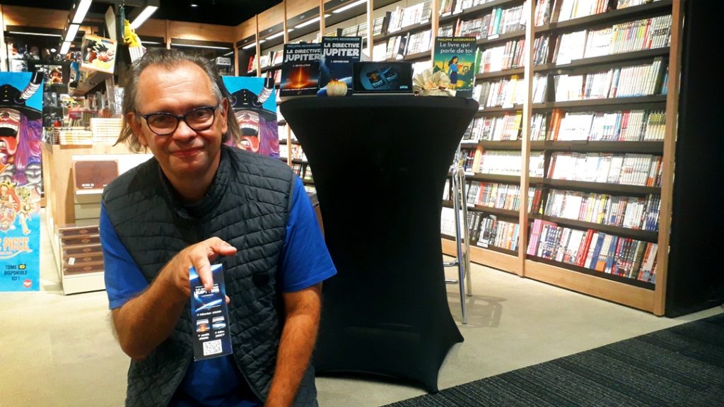 Philippe Meisburger posant avec son marque-page devant sa table de dédicace, au Leclerc d'Issenheim - 26 septembre 2026